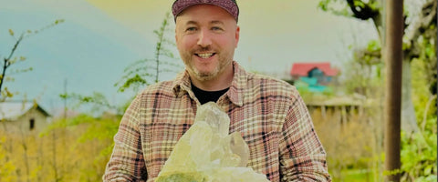 Man holding a large crystal in a natural setting with mountains and trees in the background
